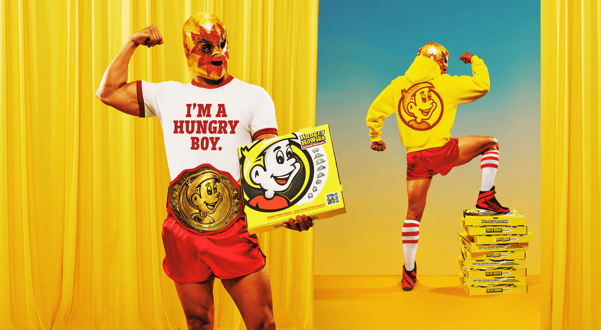 A stylized promotional scene with two people in bright yellow and red outfits posing in front of yellow curtains. One person holds a large yellow pizza box featuring a cartoon character and a championship belt, while the other stands on a stack of the pizza boxes.
